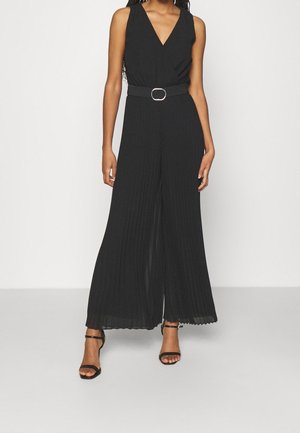 Vrouw die een mouwloze zwarte jumpsuit met ceintuur draagt, met wijde geplooide pijpen en zwarte open teen hakken, staand tegen een effen lichte achtergrond.