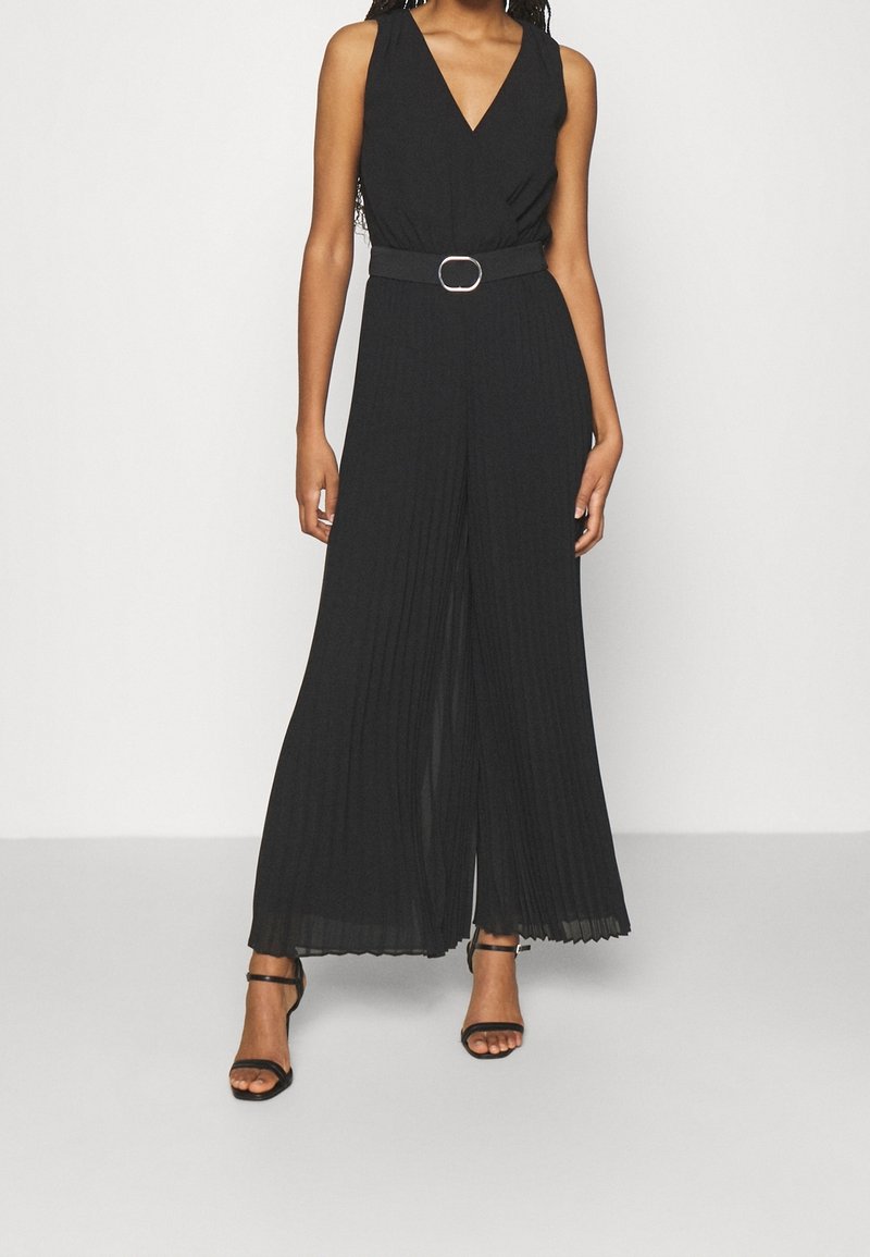 Femme portant une combinaison noire sans manches ceinturée, avec des jambes larges plissées et des talons ouverts noirs, debout devant un fond clair uni.