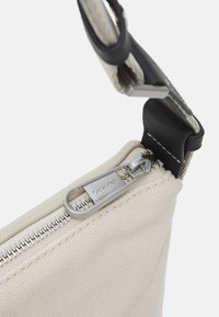Beige canvas tas met zilveren rits en leren schouderband. Heeft een opvallende ritslus met "Calvin Klein" gegraveerd. Gladde textuur.