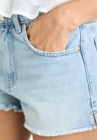 Svetlo modri jeans kratki hlače z izvlečenim robom, z bočno žepom in bronastim gumbom kot okras. Tkanina se zdi mehka in obrabljena.