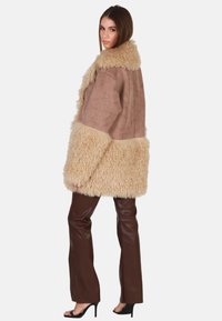 Bruine faux-suède buitenjas met een fluffy crèmekleurige bontafwerking. Met brede mouwen en een losse pasvorm, gecombineerd met bruin leren broeken.