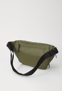 Bolsa de tela acolchada en color verde oliva, de forma triangular, con una correa negra ajustable y un bolsillo con cremallera en la parte frontal para almacenaje.