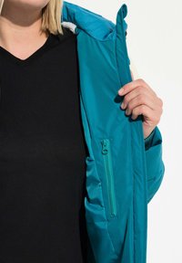 Giacca isolante color teal con tasca con zip, fodera morbida e cappuccio aderente. L'interno è foderato con un tessuto di colore contrastante.