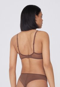 Frau mit kurzen schwarzen Haaren, die einen braunen BH mit verstellbaren Trägern und passende Unterwäsche trägt, von hinten vor einem einfarbigen Hintergrund gesehen.