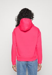 Persona con capelli ricci che indossa una felpa fucsia brillante e jeans blu, vista di schiena su uno sfondo bianco uniforme.