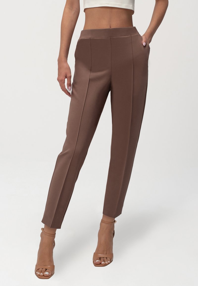 Rjave spodnje hlače iz gladkega materiala s slim fit krojem, ki imajo visok pas in sprednje gube. V kombinaciji z sandali na peto.