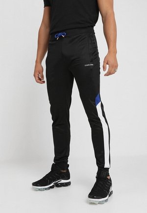 Mann trägt schwarze Sporthose mit blauen und weißen Seitenstreifen, blauer Kordel, schwarze Turnschuhe, steht vor neutralem Hintergrund.