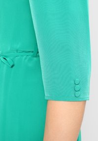 Turquoise dress sleeve featuring a smooth fabric, three fabric-covered buttons at the cuff, and a tied waist detail.