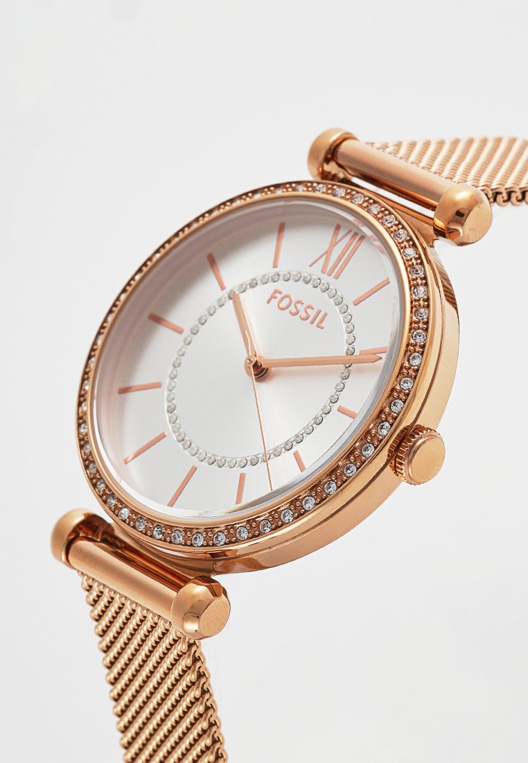 Różowe złote zegarek z białą tarczą, akcenty kryształowe wokół tarczy i siateczkowy pasek. Rzymska cyfra XII u góry.