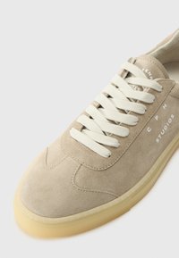 Beige suede sneaker with cream laces, rounded toe, and subtle stitching details. Features a translucent rubber sole. Brand name on the side.