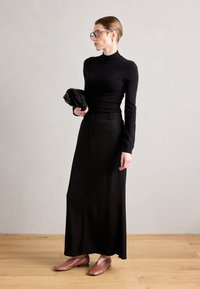 Svart långärmad åtsittande tröja med hög halsringning, i kombination med en svart maxikjol. Modellen bär bruna lagskor och håller en svart kuvertväska.