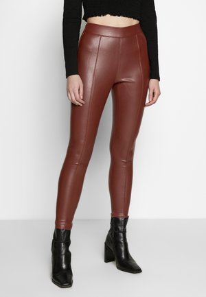 Vrouw draagt een bruine leren legging met hoge taille en zwarte enkellaarsjes, met een kort zwart topje met lange mouwen, staand tegen een witte achtergrond.