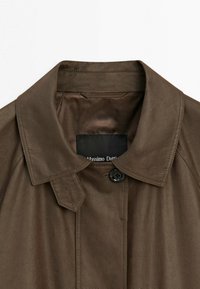 Brun frakke med en bred krave og en enkelt sort knap nær halsen, der viser syningsdetaljer og en sort stofetiket inde i kraven.