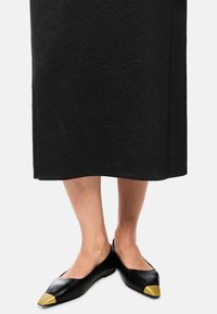 Schwarze flache Schuhe aus Leder mit spitz zulaufender Zehenpartie und einem goldfarbenen Metallkappen-Akzent. Das Design hat eine glatte Textur und minimale Nähte.