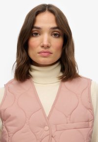 Gilet matelassé rose avec un col en V, fermeture à boutons et une petite poche sur la poitrine, superposé sur un pull-over beige à col roulé. Texture lisse.