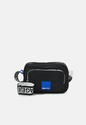 Sac bandoulière rectangulaire noir avec poche avant, écusson logo bleu et sangle blanche ajustable avec textes noirs.