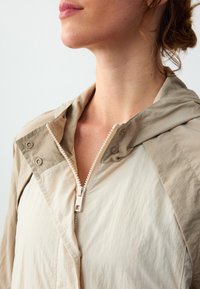 Leichte Jacke in Creme- und Taupe-Tönen, mit Reißverschluss, Druckknöpfen und einer Kapuze mit strukturierter Oberfläche des Stoffes.