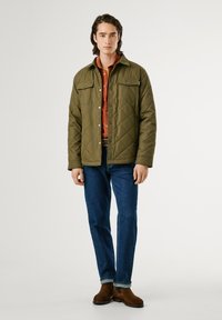 Olijfgroene doorgestikte jas met een voorste drukknoopsluiting en twee borstzakken, gedragen over een roestkleurig shirt, in combinatie met blauwe jeans en bruine laarzen.