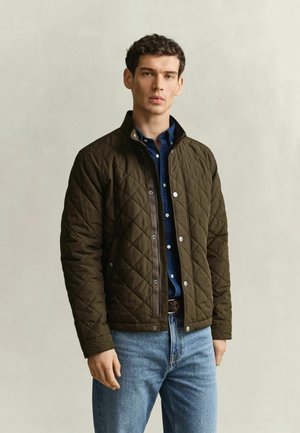 Junger Mann trägt eine dunkel olivgrüne gesteppte Jacke über einem blauen Hemd mit Knöpfen und hellblauen Jeans, steht vor einem einfarbigen hellen Hintergrund.