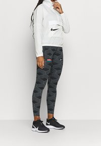 Veste blanche zippée avec une poche avant, associée à un legging noir à motifs hexagonaux comprenant un patch du drapeau du Kenya et des chaussures de sport noires.