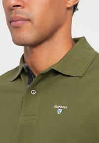 Olivově zelená polo košile s texturovanou tkaninou, límečkem a modrým kostkovaným akcentem uvnitř límce. Vybavena stříbrným knoflíkem a vyšitým logem.