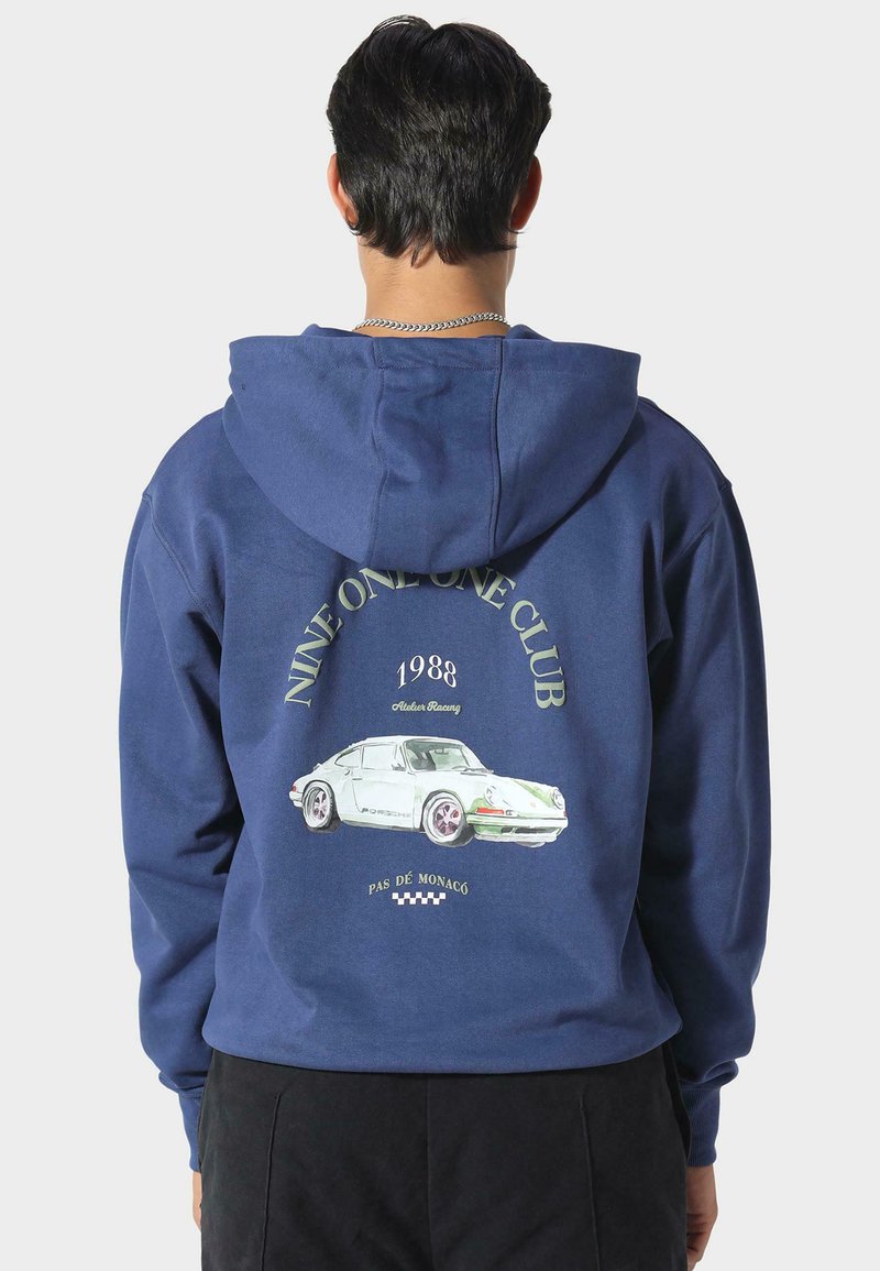 Felpa blu navy con cappuccio, caratterizzata da una grafica di una Porsche bianca sul retro, con il testo "NINE ONE ONE CLUB" e "1988" sopra. Materiale in cotone.