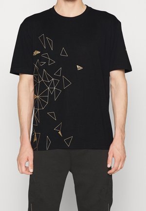 Czarny t-shirt z krótkim rękawem, ozdobiony złotymi geometrycznymi wzorami trójkątnych linii po lewej stronie, noszony przez stojącą osobę.