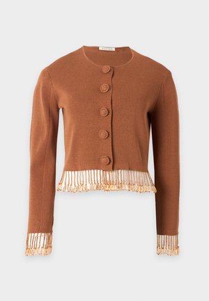 Kvinde iført mørke solbriller, brun knappet kort cardigan med frynser, lang gennemsigtig hvid nederdel og sorte flade sko, stående med hænderne nedad.