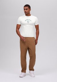 Camiseta blanca de manga corta con un gráfico de "BROOKLYN NY", combinada con pantalones beige y zapatillas blancas. Textura suave, corte casual.