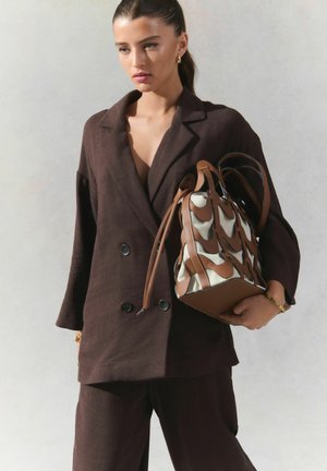 Blazer double boutonnage texturé marron avec décolleté en V, associé à un pantalon assorti. Le modèle tient un sac à main à motif marron et crème.