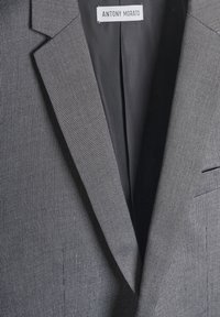 Sivi blejzer s teksturiranim materijalom, s nazubljenim reverima i jednom džepom. Oznaka glasi "ANTONY MORATO." Unutrašnja podstava čini se da je crna.