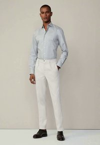 Man wearing light blue striped dress shirt, white trousers, and black dress shoes, standing against a plain beige background.