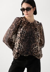 Blouse à imprimé léopard marron et noir avec des manches longues et transparentes, un col arrondi et une ouverture en goutte. Associée à un jean noir et des lunettes de soleil.