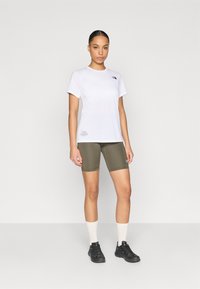 Λευκό κοντομάνικο μπλουζάκι με μαύρο λογότυπο, συνδυασμένο με αθλητικό σορτς σε ελιά πράσινο χρώμα. Αξεσουάρ περιλαμβάνουν λευκές κάλτσες και μαύρα αθλητικά παπούτσια.