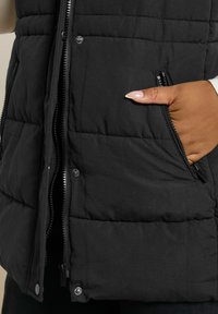 Svart vadderad väst med blixtlås framtill, tryckknappar och sidofickor. Materialet är tjockt och texturerat med horisontell sömnad.