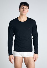 Juoda ilgomis rankovėmis marlė t-shirts, pagaminta iš medvilnės mišinio, su apvaliu iškirpimu ir mažuoju baltu "BOSS" logotipu krūtinės srityje. Dėvima su baltomis apatinėmis kelnaitėmis.