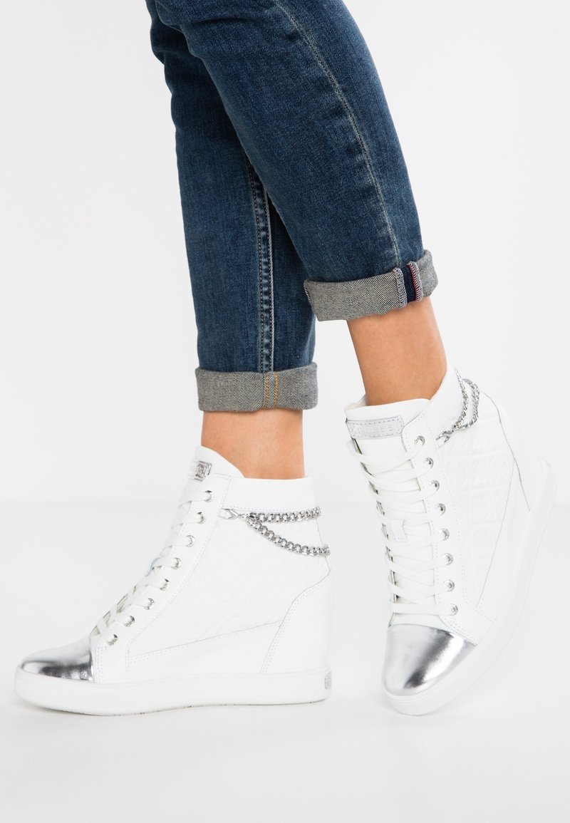 Des baskets montantes blanches présentent un côté texturé, un embout en métal argenté et une chaîne décorative sur le dessus. Elles sont associées à un jean roulé.