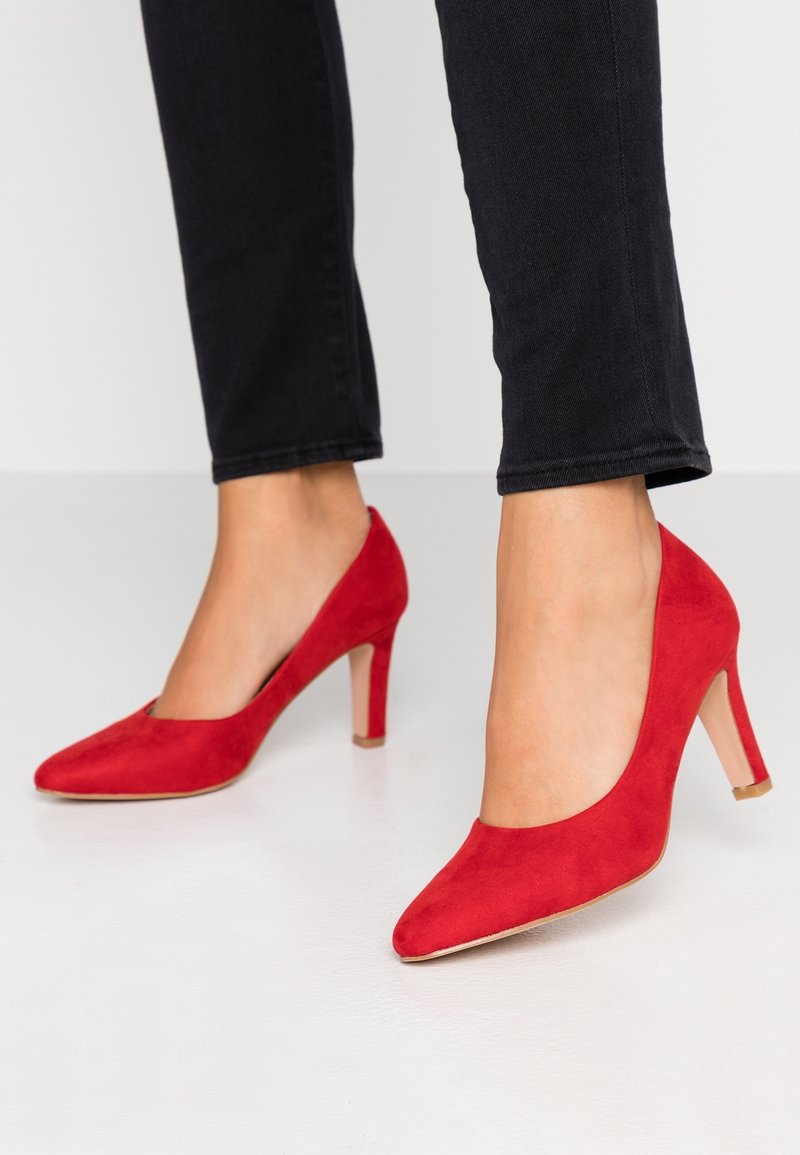 Escarpins en daim rouge avec un bout pointu et un talon modeste. La chaussure présente une texture lisse et un style ajouré sur les côtés.