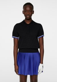 Svart polo skjorta med präglat mönster, korta ärmar med blå och vit kant, i kombination med en plisserad blå kjol och en vit handske.