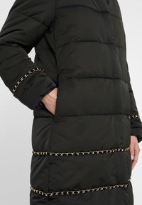 Czarna pikowana kurtka z poziomym przeszyciem, bocznymi kieszeniami oraz ozdobnymi złotymi łańcuszkami na rękawach i tułowiu. Miękka, gładka faktura.