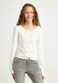 Hvid langærmet top med snørelukning foran, med nitter og ribbet tekstur. Kombineret med lysegrå jeans.