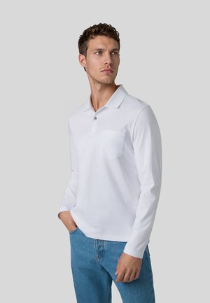 Junger Mann mit kurzen lockigen Haaren, der ein weißes Poloshirt mit langen Ärmeln und blaue Jeans trägt, steht mit einer Hand in der Tasche und schaut zur Seite.