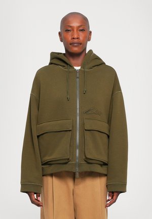 Personne portant une veste à capuche zippée vert olive avec de grandes poches avant et un pantalon beige, se tenant contre un fond blanc.
