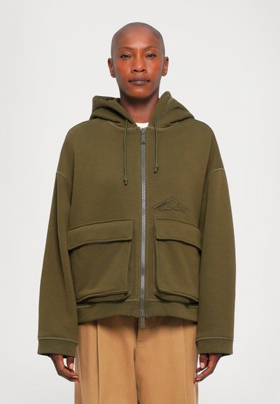 Personne portant une veste à capuche zippée vert olive avec de grandes poches avant et un pantalon beige, se tenant contre un fond blanc.
