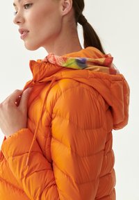 Giacca arancione imbottita con cappuccio a fantasia colorata, dotata di chiusura con zip e regolazioni con cordino. Design strutturato e voluminoso.