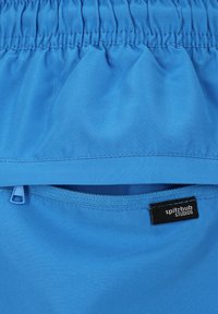 Blauwe stof met een soepele textuur, voorzien van een elastische tailleband en een ritszak. Een label met de tekst "spitzbub STUDIOS" bevindt zich nabij de zoom.