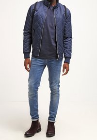 Navy quilted bomber jacket with a high collar, worn over a textured navy sweater. Slim-fit blue jeans and dark brown ankle boots complete the look.