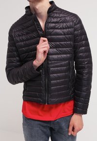 Svart fyrkantig jacka med ståkrage, dragkedjestängning och två framfickor. Bärs över en röd skjorta. Lätt material och figurnära design.