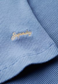 Tejido de rayas azul con un acabado texturizado; presenta un logotipo bordado en dorado "Superdry" en el borde.