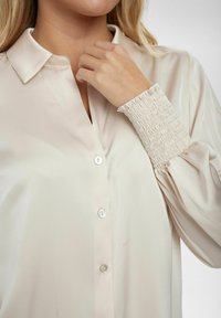 Blusa beige con scollo a colletto, tessuto di raso liscio, chiusura frontale a sei bottoni e texture arricciata ai polsini.